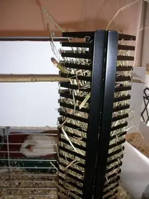 Eine graue Heuraufe aus Metall steht in einem Käfig und ist mit Heu gefüllt, während ein Meerschweinchen im Hintergrund sitzt.