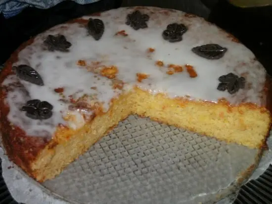 Ein saftiger Orangen-Mandelkuchen, glasiert und mit dekorativen Blättern, steht auf einem Kuchengitter in einer gemütlichen Küche.