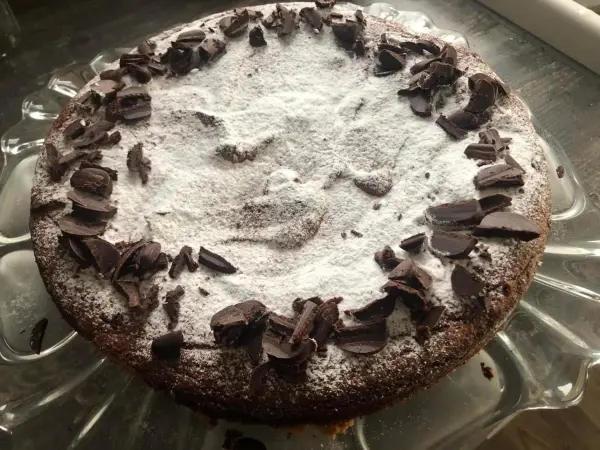 Ein runder Orangen-Mandelkuchen liegt auf einem gläsernen Teller, dekoriert mit Puderzucker und Schokoladenspänen.