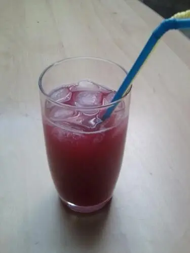 Ein Glas mit rotem Rhabarber-Eistee steht auf einem Holztisch, ein bunter Strohhalm ragt heraus.