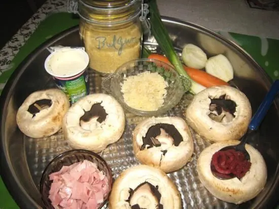 Auf einem Tablett liegen halbierte Champignonköpfe, bereit zum Füllen mit Bulgur, Schinken und Creme fraiche, umgeben von frischen Zutaten.