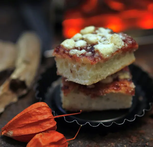 Zwei Stücke Zwetschgenkuchen mit Streuseln liegen übereinander auf einer dunklen Unterlage, umgeben von orangefarbenen Dekorationen.