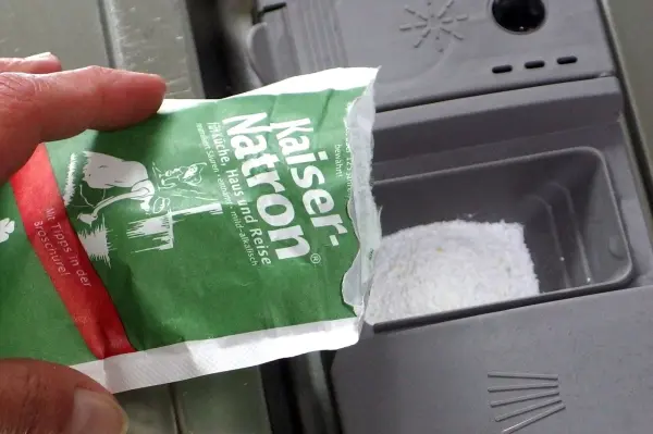 Ein Hand hält ein Päckchen Speisenatron über das Fach einer Spülmaschine, bereit für die Anwendung zur Reinigung.