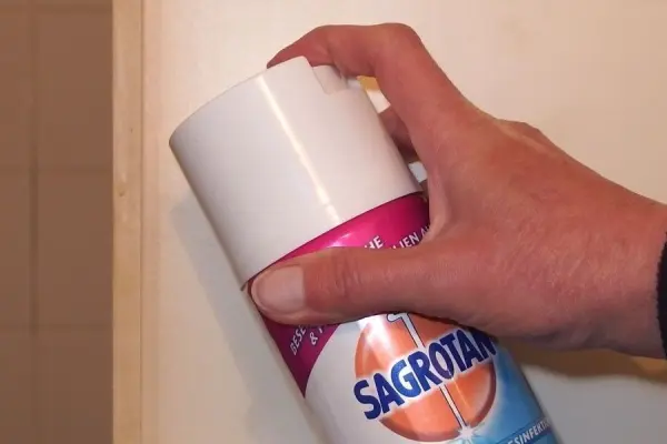 Ein Hand hält eine Sagrotan-Sprühflasche, um Klebereste auf Kunststoff zu entfernen, während sich eine Wand im Hintergrund befindet.