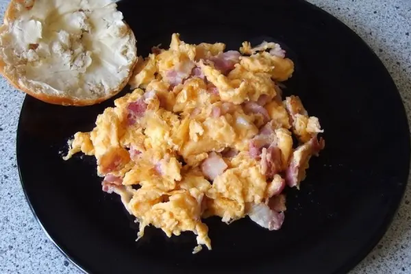 Lecker Rührei liegt auf einem schwarzen Teller, während es neben einem halben Brötchen in einer einfachen Küche präsentiert wird.