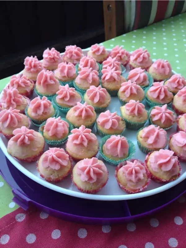 Bunte Cupcakes stehen auf einem weißen Teller, dekoriert mit rosa, veganem Frosting, umgeben von einem bunte Tischdecke.