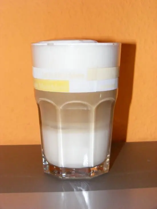 Ein zweifarbiges Latte Macchiato füllt ein Glas, während es in einem warmen Raum vor orangefarbener Wand steht.