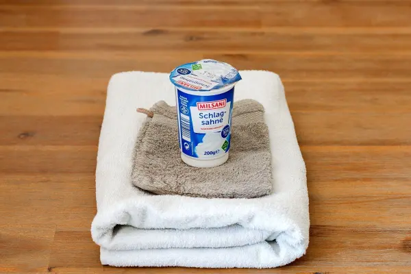 Ein Becher Milsani Schlagsahne steht auf einem hellen Handtuch und einem Waschhandschuh auf einem Holztisch.
