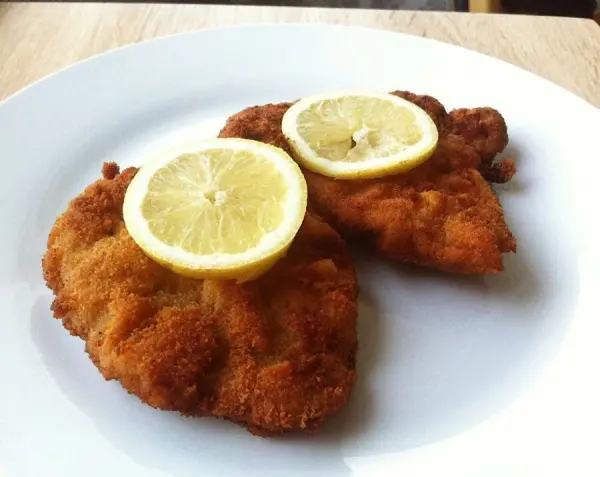 Zwei knusprige Schnitzel auf einem weißen Teller, garniert mit Zitronenscheiben, an einem hellen Holzuntergrund.