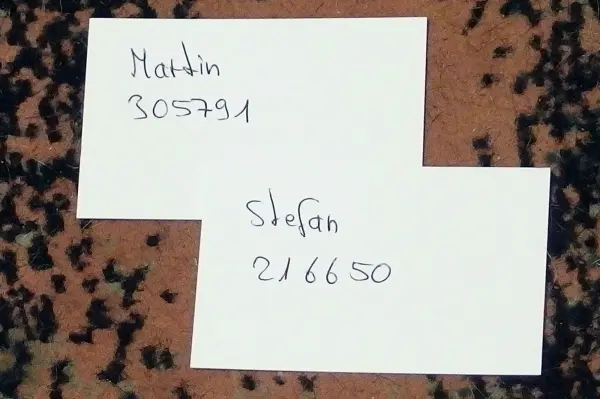 Kleine Zettel liegen auf einem Teppich, auf denen Namen und Zahlen handgeschrieben sind: „Martin 305791“ und „Stefan 216650“.