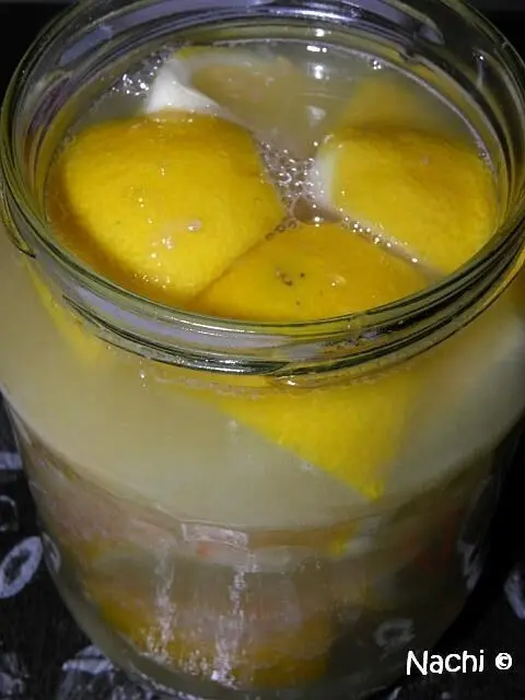 Ein Glas mit eingelegten Zitronen schwimmt in einer transparenten Flüssigkeit, umgeben von einer dunklen Oberfläche.