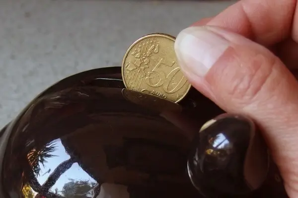 Eine Hand steckt eine Münze in ein braunes Sparschwein, um Kindern den Umgang mit Geld näher zu bringen.