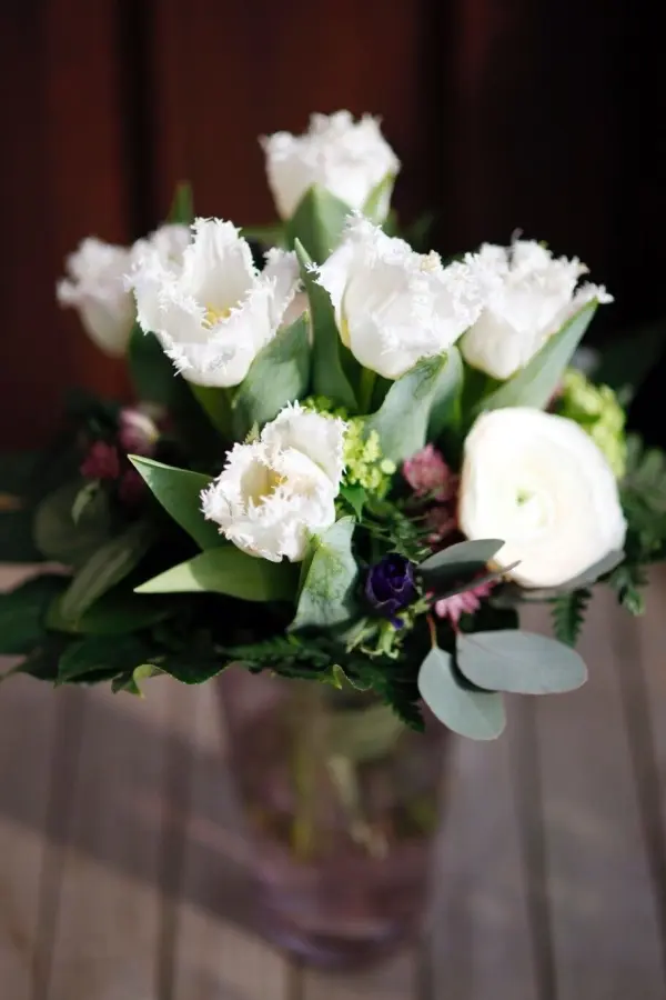 Ein Blumenstrauß mit frischen weißen Tulpen steht in einer Glasvase auf einem Holztisch.