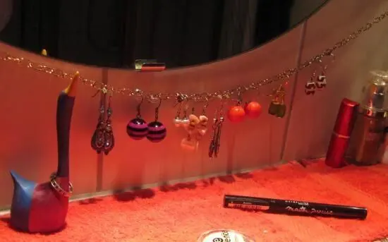 Ohrringe hängen an einer Kette über einem Badspiegel, während ein roter Schminkuntergrund und Kosmetikartikel sichtbar sind.