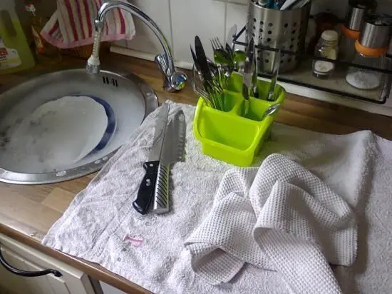 Auf einer Küchentheke liegt ein Handtuch, während Geschirrspülen im Hintergrund mit einem Waschbecken und Besteckhalter passiert.