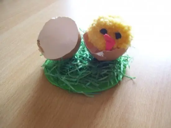 Ein gelbes, flauschiges Küken sitzt auf grünem Gras und schaut aus einer zerbrochenen Eierschale, fröhliche Osterdeko.