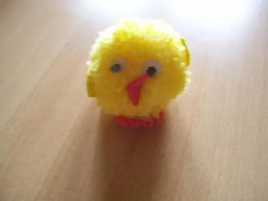 Ein gelbes Pomponküken sitzt auf einem Tisch, seine runden Augen schauen fröhlich hervor, perfekt für Osterdeko.