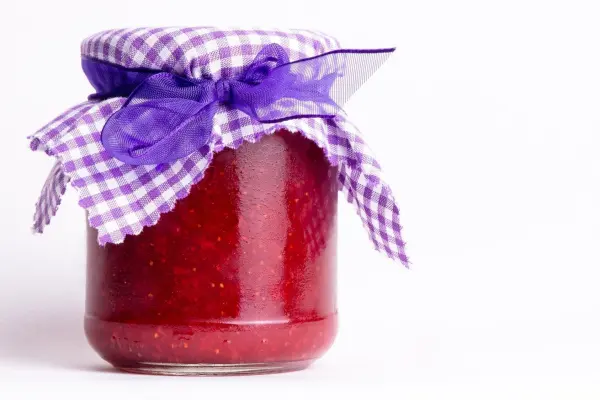 Ein Glas mit roter Marmelade steht auf weißem Hintergrund, bedeckt mit einem lilafarbenen karrierten Stoff und Schleife.
