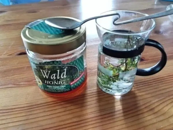 Ein Glas mit Thymiantee steht neben einem Honigglas, während ein Löffel auf dem Glas ruht. Holzoberfläche als Hintergrund.