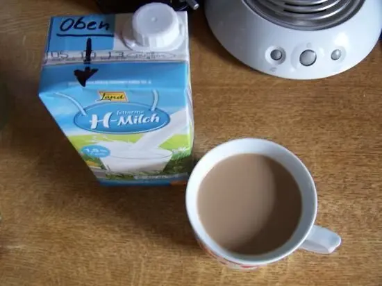 Ein Tetra Pak Milch steht neben einer Tasse mit braunem Kaffee auf einem Tisch, während ein Wasserkocher im Hintergrund sichtbar ist.