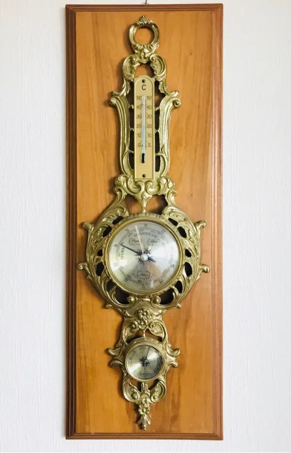 Ein antikes Thermometer und Barometer hängen an einer Holzplatte, die die Raumtemperatur und den Luftdruck anzeigt.