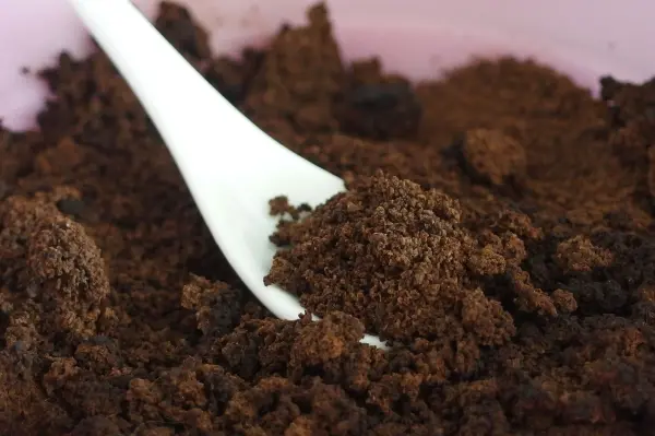 Ein weißer Löffel greift in ein Schälchen mit dunklem Kaffeepeeling, das auf einem rosa Untergrund liegt.