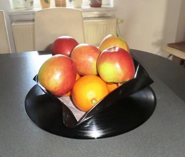 Eine Obstschale aus Schallplatte hält mehrere Äpfel und eine Orange auf einem Tisch in einem hellen Raum.