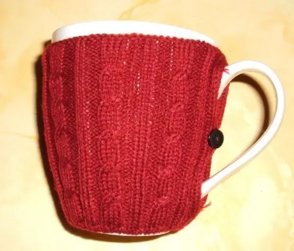 Eine rote Strickhülle umschließt eine weiße Kaffeetasse, die auf einem hellen Untergrund steht und Wärme spendet.