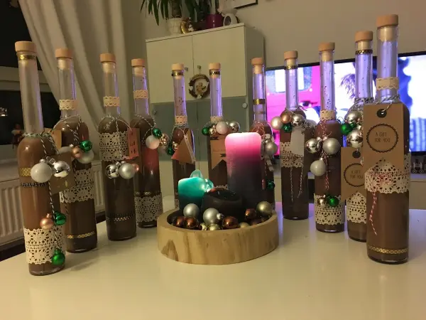 Flaschen mit Lebkuchen-Likör stehen dekoriert mit Weihnachtskugeln und Spitzenband auf einem Tisch, umgeben von Kerzen.