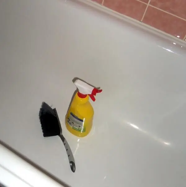 Ein gelber Sprühflakon und Handfeger liegen in einer weißen Badewanne mit rosa Fliesen im Hintergrund.
