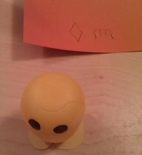 Ein gelbes Überraschungsei-Küken mit schwarzen Augen steht auf einem Tisch, während ein orangefarbener Zettel dahinter liegt.