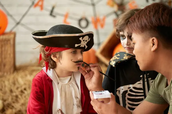 Ein Kind im Piratenkostüm sitzt ruhig, während jemand ihm einen Schnurrbart aufmalt, umgeben von festlicher Dekoration.