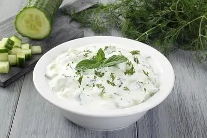 Eine weiße Schale mit Tzatziki aus Quark und Gurkenwürfeln auf einem Holztisch, garniert mit Dill und Minze, umgeben von frischen Kräutern.