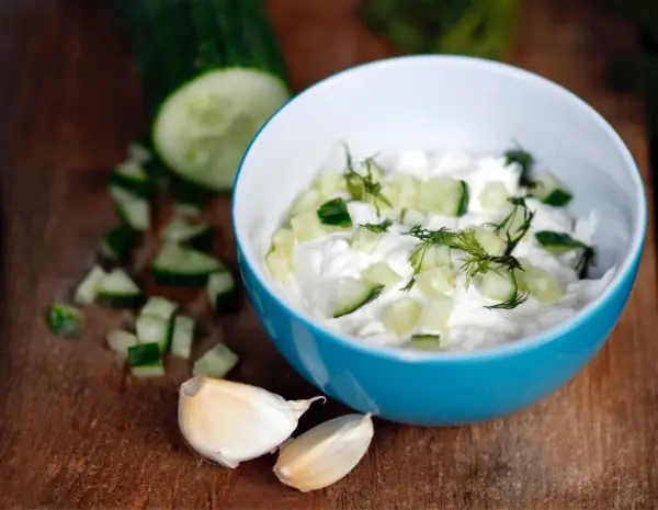 Eine Schüssel mit Tzatziki aus Quark und Gurkenstückchen steht auf einem Holzbrett, umgeben von gehacktem Knoblauch.