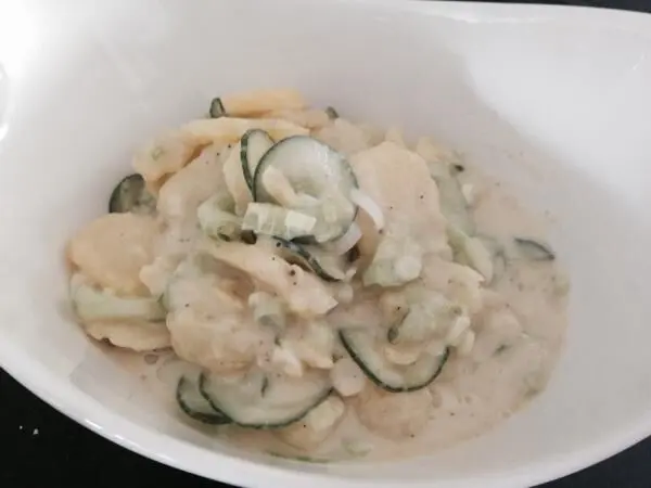Kartoffelsalat mit Gurken liegt in einer weißen Schüssel; die Zutaten sind mit einer cremigen Sauce vermischt.