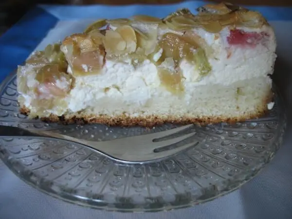 Eine fruchtige Quark-Rhabarber-Torte mit Mandelbaiser liegt auf einem transparenten Teller und wird mit einer Gabel serviert.