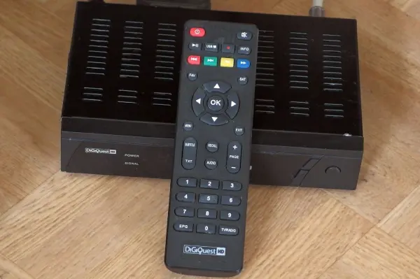 Eine TV-Fernbedienung liegt auf einem SAT-Receiver auf einem Holzfußboden, bereit zur Nutzung ohne direkten Kontakt.