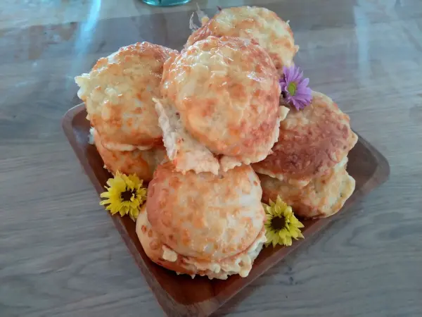 Auf einem Holzbrett liegen goldbraune Käsebrötchen, umgeben von bunten Blumen. Sie wirken frisch und einladend.