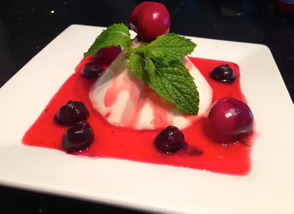 Eine Panna Cotta in Vanilleweiß sitzt auf roter Erdbeersauce, garniert mit frischen Minzblättern und umgeben von Kirschen.