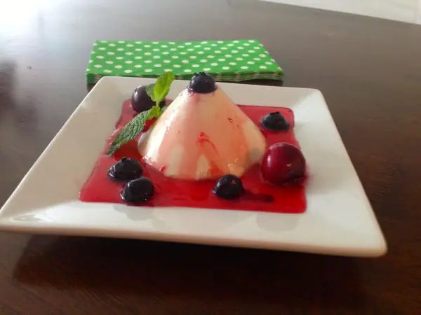 Panna Cotta ruht auf weißem Teller mit roter Soße und Beeren, dekoriert mit Minzblättern; grüne Serviette im Hintergrund.