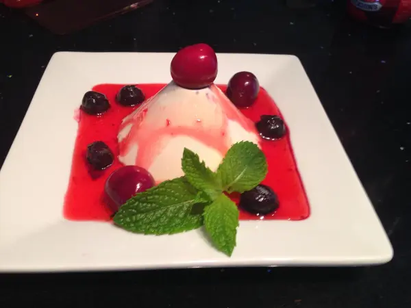 Panna Cotta mit roten Früchten und Minzblättern auf weißem Teller, umgeben von roter Sauce auf dunklem Untergrund.
