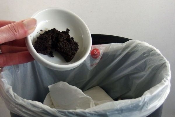 Eine Hand hält eine kleine Schüssel mit etwas Müll über einen Mülleimer, in dem sich bereits Abfälle befinden.