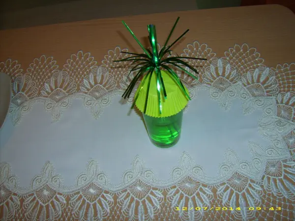 Ein grüner Becher mit gelber Abdeckung und einer Spinne aus Plastik schützt Getränke vor Insekten, auf einem weißen Tischläufer.
