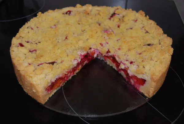 Eine Zwetschgen-Streusel-Torte mit krümeliger Streußelschicht, die teilweise angeschnitten auf einer schwarzen Oberfläche liegt.