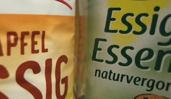 Zwei Flaschen Essig stehen nebeneinander, eine für Apfelessig, die andere für Essigessenz, beide in heller Umgebung.