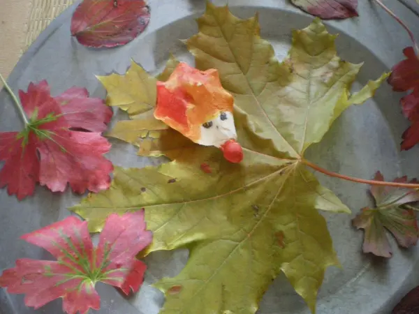 Ein kleiner Salzteig-Igel liegt auf einem großen, grünen Ahornblatt, umgeben von roten Herbstblättern auf einem Metalltablett.