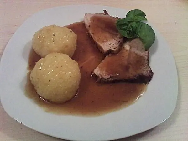 Zartes Schweinebratenstück liegt auf einem Teller, umgeben von zwei Kartoffelknödeln in reichhaltiger Sauce.