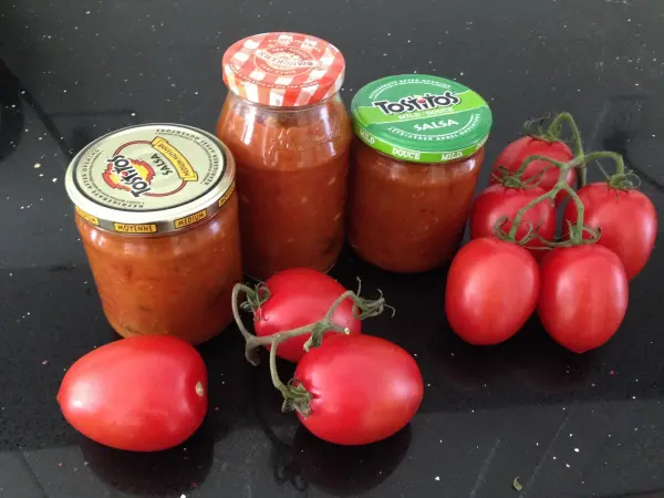 Glänzende rote Tomaten liegen neben drei Gläsern mit Tomaten-Chutney auf einer dunklen Oberfläche, bereit zum Kochen.