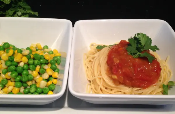 Gekochte Spaghetti mit Tomaten-Chutney und frischem Koriander, neben einem bunten Salat aus Erbsen und Mais serviert.