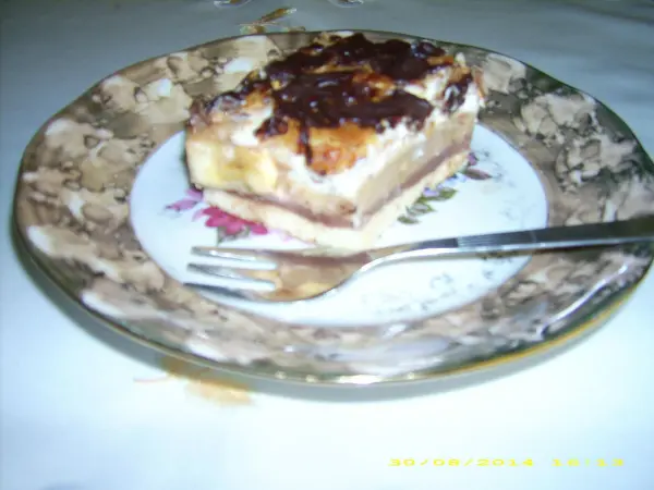 Ein Stück Schoko-Karamell-Bananenkuchen liegt auf einem blumengemusterten Teller mit einer Gabel daneben, umgeben von hellem Tisch.